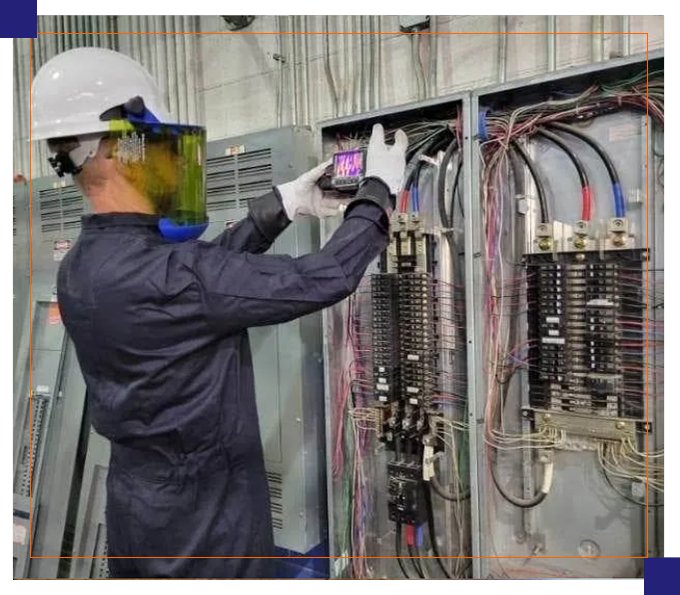 A man in white helmet and blue coveralls working on electrical equipment.